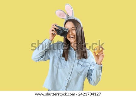 Young woman in bunny ears with photo camera and Easter egg on yellow background