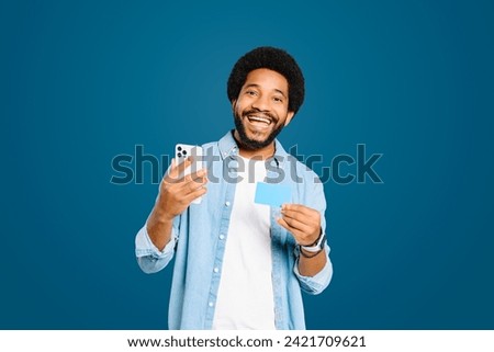 Excited Brazilian man holds up a smartphone in one hand and a credit card in the other, ideal for concepts related to online shopping or mobile banking standing isolated on blue, e-banking concept