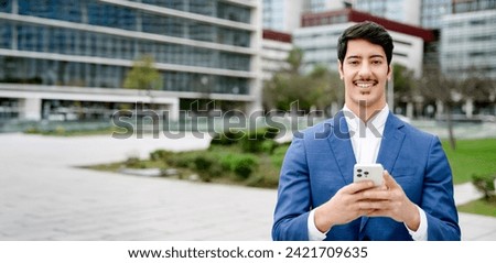 With a wide cityscape stretching behind him, a Hispanic entrepreneur smiles at the camera while using his smartphone, symbolizing the agile nature of contemporary business.