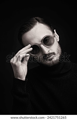 Art photography. Portrait of a brutal confident dark-haired man in round glasses standing in the dark on a black background. A beam of light illuminates his face.