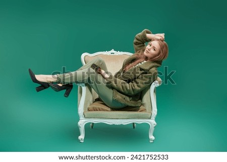 Full-length portrait of a stylish fashion model girl posing in vintage armchair in green trousers and green sheepskin coat. Fashion photo. 
