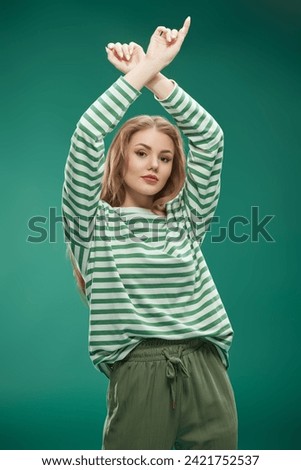 A joyful girl in green trousers and green striped longsleeve dancing and having fun. Studio portrait on a green background. St. Patrick's Day celebration.
