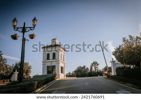 Sotogrante, Spain - January 27, 2024 -  street view with a white tower-like building with a tiled roof and arched windows, a streetlamp with hanging flower pots, and lush trees.