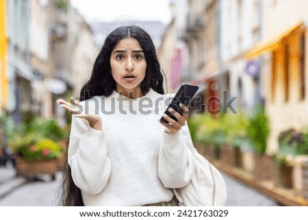 Portrait of an Indian serious and shocked woman standing in the city on the street, holding the phone in her hands and looking worriedly at the camera. Royalty-Free Stock Photo #2421763029