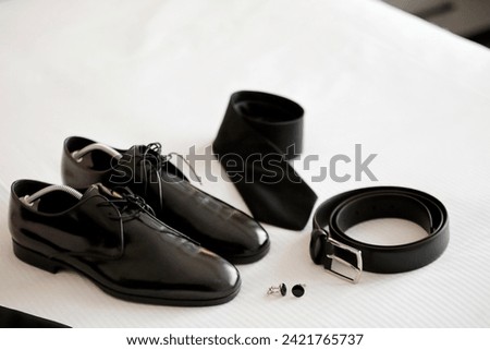In the groom's room, a white sheet covers the bed as black patent leather shoes, a tie, a belt, and cufflinks lay neatly arranged, capturing a close-up composition of wedding day essentials. stock pho