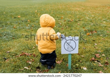 Little cute baby toddler child in yellow jacket walks through puddles in the autumn park stands near the lawn with a sign forbidding walking on the lawn