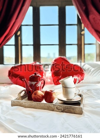 Two white cups, two ripe apples and a red coffee pot and two heart-shaped balloons in blurry focus are on a white bed lit by bright morning light in vintage interior. Vertical image. Valentine’s Day.