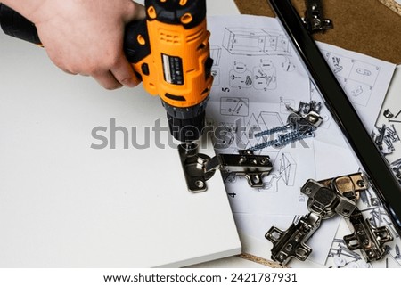 Installing a furniture hinge on the door. Assembling furniture and fixing fittings close-up. Screwing the door hinge onto a piece of furniture with a screwdriver. Royalty-Free Stock Photo #2421787931