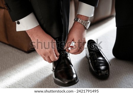 The groom, dressed in a tuxedo, leans down to tie his patent leather shoes in a close-up shot captured in the groom's room. stock photo