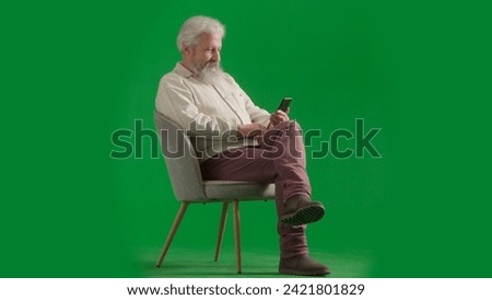 Portrait of aged bearded man on chroma key green screen background. Full shot of senior man sitting on a chair holding smartphone, reading news.