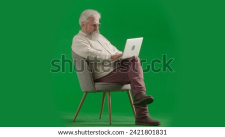 Portrait of aged bearded man on chroma key green screen background. Full shot of senior man sitting on a chair working on laptop.