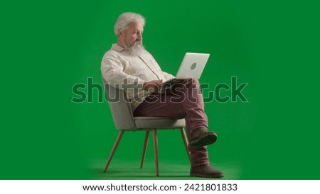 Portrait of aged bearded man on chroma key green screen background. Full shot of senior man sitting on a chair watching video on laptop.
