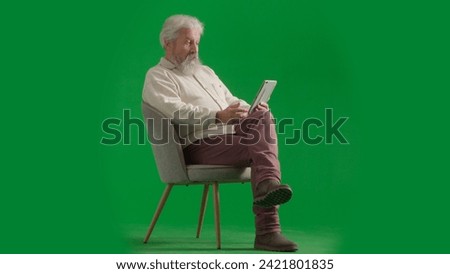 Portrait of aged bearded man on chroma key green screen background. Full shot of senior man sitting on a chair holding tablet watching media.