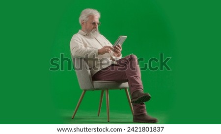 Portrait of aged bearded man on chroma key green screen background. Full shot of senior man sitting on a chair watching video on tablet.