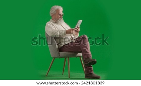 Portrait of aged bearded man on chroma key green screen background. Full shot of senior man sitting on a chair reading book on tablet.