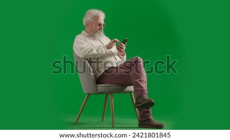 Portrait of aged bearded man on chroma key green screen background. Full shot of senior man sitting on a chair holding smartphone texting.