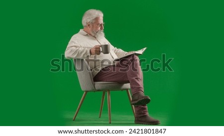 Portrait of aged bearded man on chroma key green screen background. Full shot of senior man sitting on a chair drinking tea reading book.