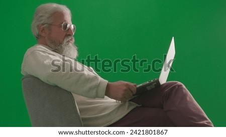 Portrait of aged bearded man on chroma key green screen background. Full shot of senior man sitting on a chair working on laptop.