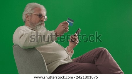 Portrait of aged bearded man on chroma key green screen background. Full shot of senior man sitting on a chair holding card and smartphone.