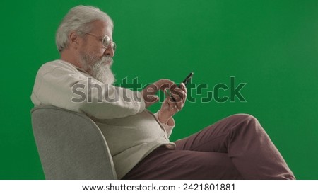 Portrait of aged bearded man on chroma key green screen background. Full shot of senior man sitting on a chair holding smartphone texting.