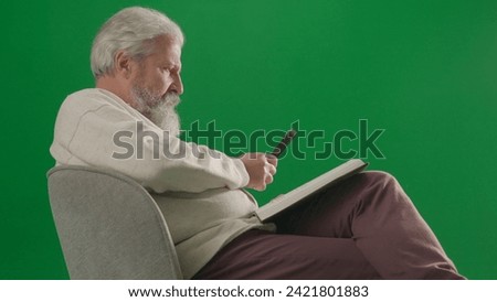 Portrait of aged bearded man on chroma key green screen background. Full shot of senior man sitting on chair reading book with magnifying glass.