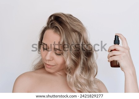 Young caucasian attractive blonde woman with wavy hair spraying thermal protection hairspray, oils with vitamins in a brown bottle on a light background Royalty-Free Stock Photo #2421829277