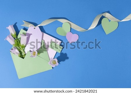 Top view of an envelope in pastel green color filled with some flowers and a small paper with copy space. Paper hearts arranged with ribbon over blue background. Happy Women’s Day celebration