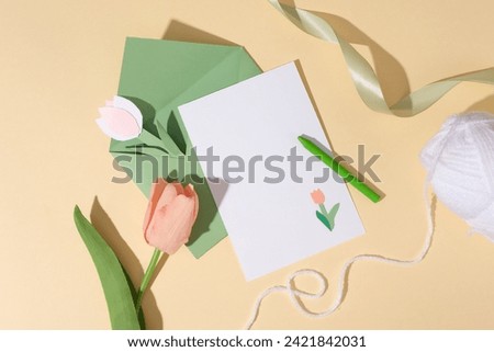 Flat lay of a white paper and green envelope decorated with a crayon, ribbon and a white skein. A tulip flower and paper tulip flowers featured. Copy space on the card for content about Women’s Day