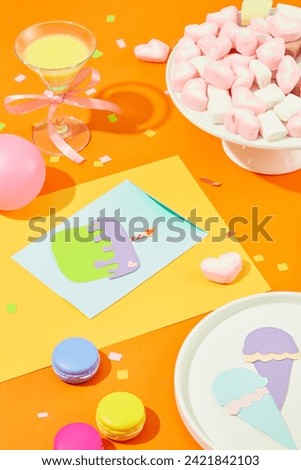 A plate of marshmallows, a cocktail, Ice cream and a birthday cake cut from colored paper, balloons and macarons on an orange background.