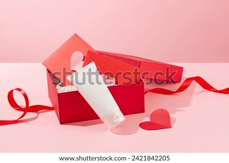 An opened gift box in red color containing few special card inside. Cosmetic tube displayed on pastel pink surface with a red ribbon. Empty label for beauty product branding mockup
