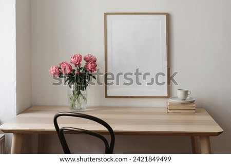 Elegant interior still life. Blank vertical picture frame mockup. Vase with pink peonies flowers. Cup of tea, coffee on books. Wooden table, desk. Romantic home breakfast, white wall background.