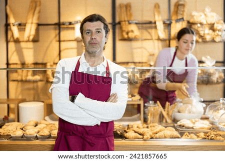 Confident middle-aged male business owner, worker in apron standing in front of pastry stand in bakery shop. Concept of small local business Royalty-Free Stock Photo #2421857665