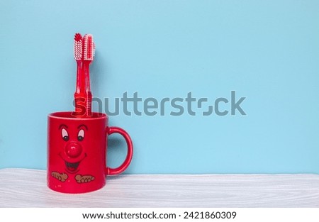 Red toothbrush in red mug cartoon face with copyspace isolated on blue background. Wallpaper red mug and toothbrush on gray table and blue background Royalty-Free Stock Photo #2421860309