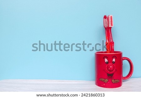 Red toothbrush in red mug cartoon face with copyspace isolated on blue background. Wallpaper red mug and toothbrush on gray table and blue background Royalty-Free Stock Photo #2421860313