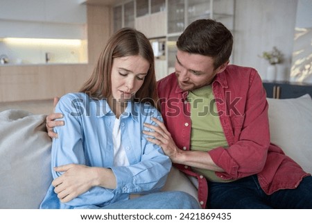 Tender caring husband sitting on sofa, calming down upset depressed wife in grief, sorrow. Loving man embracing sad female. Emotional support, consolation in difficult situations in family relations Royalty-Free Stock Photo #2421869435