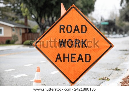 A road work ahead plastic orange sign on a side of road telling car drivers, bikers and pedestrians to be alert as they are entering a construction area and utility works with an orange cone nearby