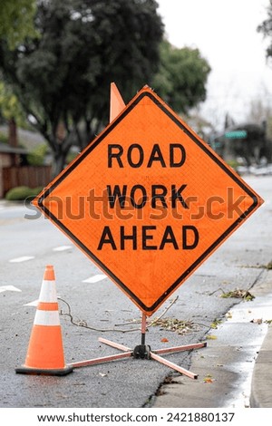 A road work ahead plastic orange sign on a side of road telling car drivers, bikers and pedestrians to be alert as they are entering a construction area and utility works with an orange cone nearby Royalty-Free Stock Photo #2421880137