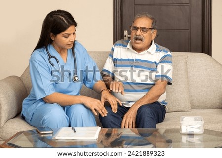 Trustful nurse checking senior male joint knee pain at home. Cheerful Indian female general practitioner communicating with pleasant 60s male patient, sitting together on sofa.  Royalty-Free Stock Photo #2421889323
