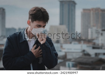 Portrait Asian young man wear N95 mask to protect PM 2.5 dust and air pollution. Young man wearing protection mask over smog city building with bad weather, pm2.5, lung cancer, City air pollution. Royalty-Free Stock Photo #2421900997