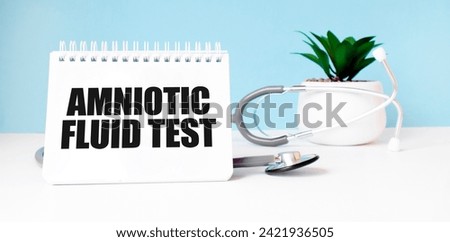 The text AMNIOTIC FLUID TEST is written on notepad near a stethoscope on a blue background. Medical concept