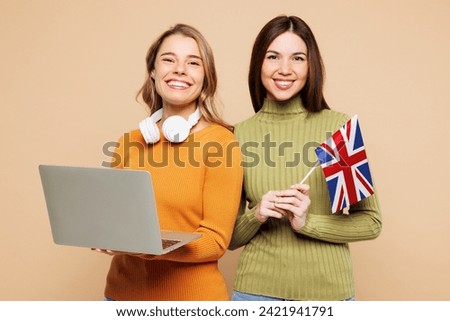 Young friends two IT women they wearing orange green shirt casual clothes together hold British flag use work on laptop pc computer isolated on plain pastel beige background studio. Lifestyle concept