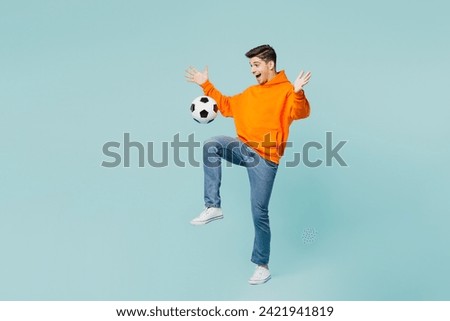 Full body excited fun cool young man fan wearing orange hoody casual clothes cheer up support football sport team hold hits soccer ball on knee watch tv live stream isolated on plain blue background