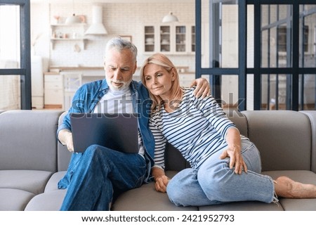 Middle age woman and man browsing internet, shopping online on notebook, sitting on sofa in living room at home. Focused on modern screen. Enjoying leisure together