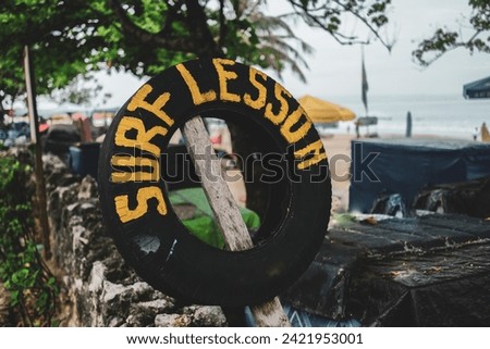 A surf lesson sign under a tree on Kuta Beach, Bali, made of recycled tires, with the Kuta Beach in the background
