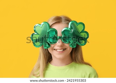 Cute little girl in decorative glasses in shape of clover on yellow background. St. Patrick's Day celebration