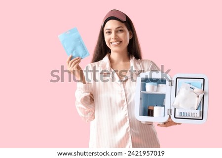 Beautiful young happy woman with facial mask and small refrigerator for cosmetic products on pink background