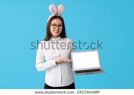 Young Asian businesswoman in bunny ears with laptop on blue background. Easter celebration