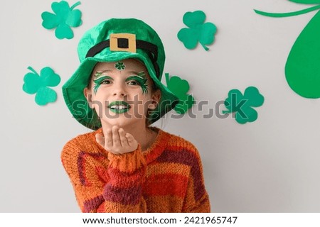 Funny girl with face painting, leprechaun's hat and clovers blowing kiss near white wall. St. Patrick's Day celebration
