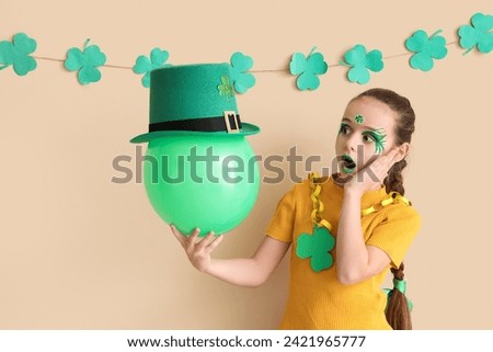 Surprised girl with face painting, balloon and leprechaun's hat near beige wall. St. Patrick's Day celebration