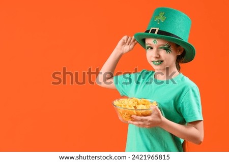 Funny girl with face painting, leprechaun's hat and potato chips on orange background. St. Patrick's Day celebration